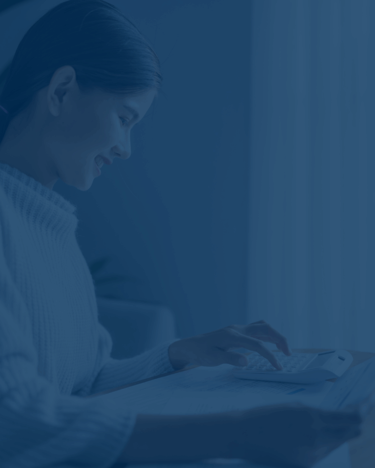 Woman in a light sweater uses a calculator on a desk with papers nearby, focused on task.