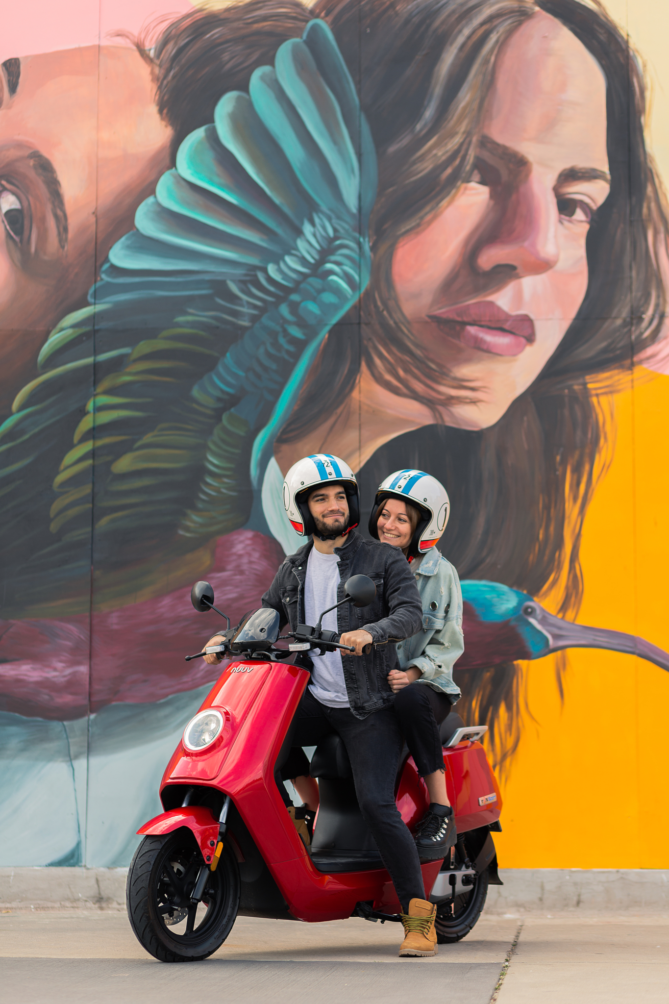 Couple wearing helmets rides a red scooter in front of a vibrant mural of a woman's face with turquoise wings.