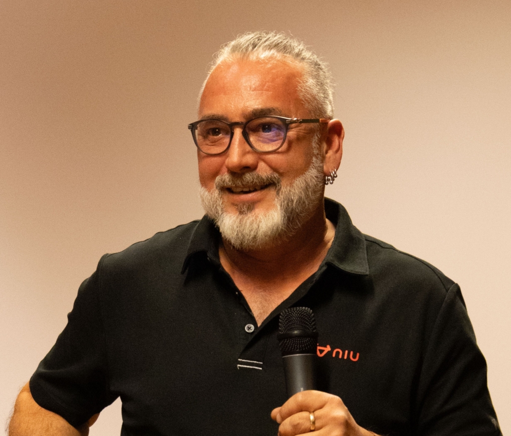 Man with gray beard and glasses, wearing a black polo, holding a microphone as he speaks on stage/at an event.